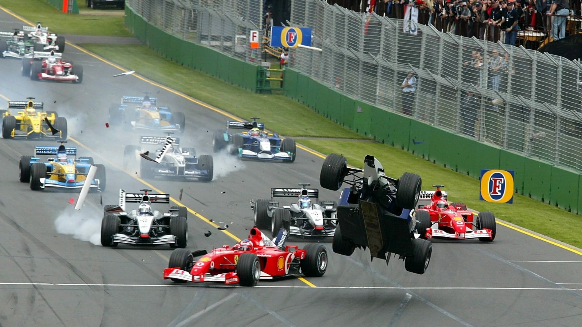 En dramatisk scen från Australiens GP 2002 där en Formel 1-bil lyfter från marken i en startkrasch, omgiven av andra bilar och rök på banan.