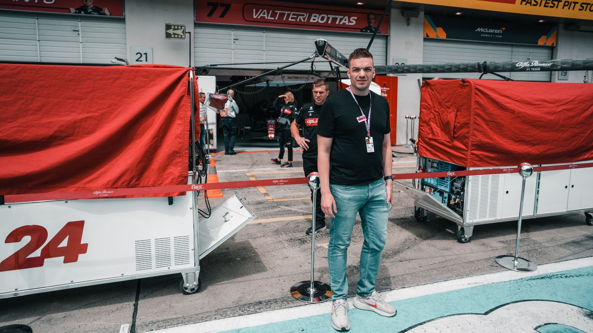 F1-reportern Christoffer Lindén framför Sauber F1-teamets garage med Valtteri Bottas namn synligt ovanför porten. Han bär en svart t-shirt, jeans och ackreditering, med garageutrustning och teammedlemmar i bakgrunden.